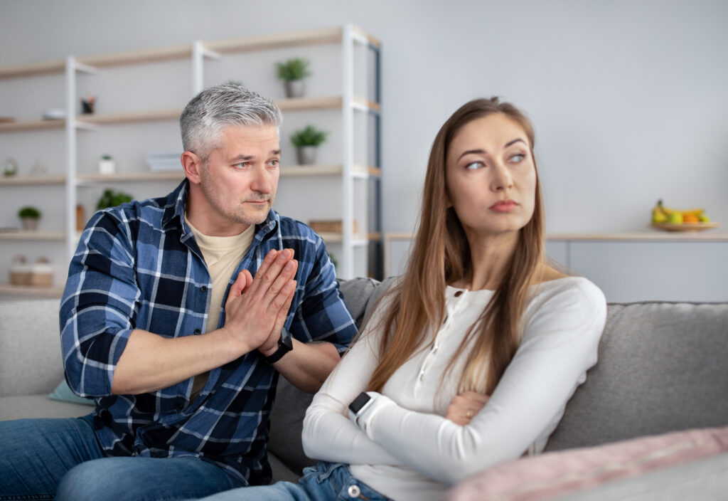 Offended mature lady sitting on couch with folded arms, her husband begging for forgiveness after fight, indoors. Senior man trying to make peace with his wife after family conflict