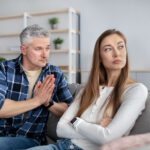 Offended mature lady sitting on couch with folded arms, her husband begging for forgiveness after fight, indoors. Senior man trying to make peace with his wife after family conflict