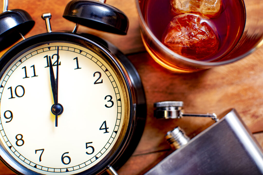 Alcoholism concept. Glass with whiskey and ice with an alarm clock and a flask on wood.