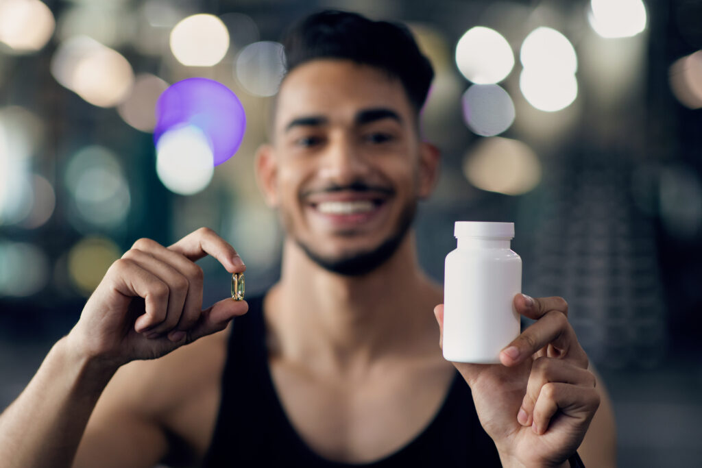 Fitness Supplements. Sporty Middle Eastern Guy Demonstrating Blank White Jar And Capsule Pill, Arab Male Athlete Advertising Multivitamins For Sports And Bisybuilding, Selective Focus, Closeup