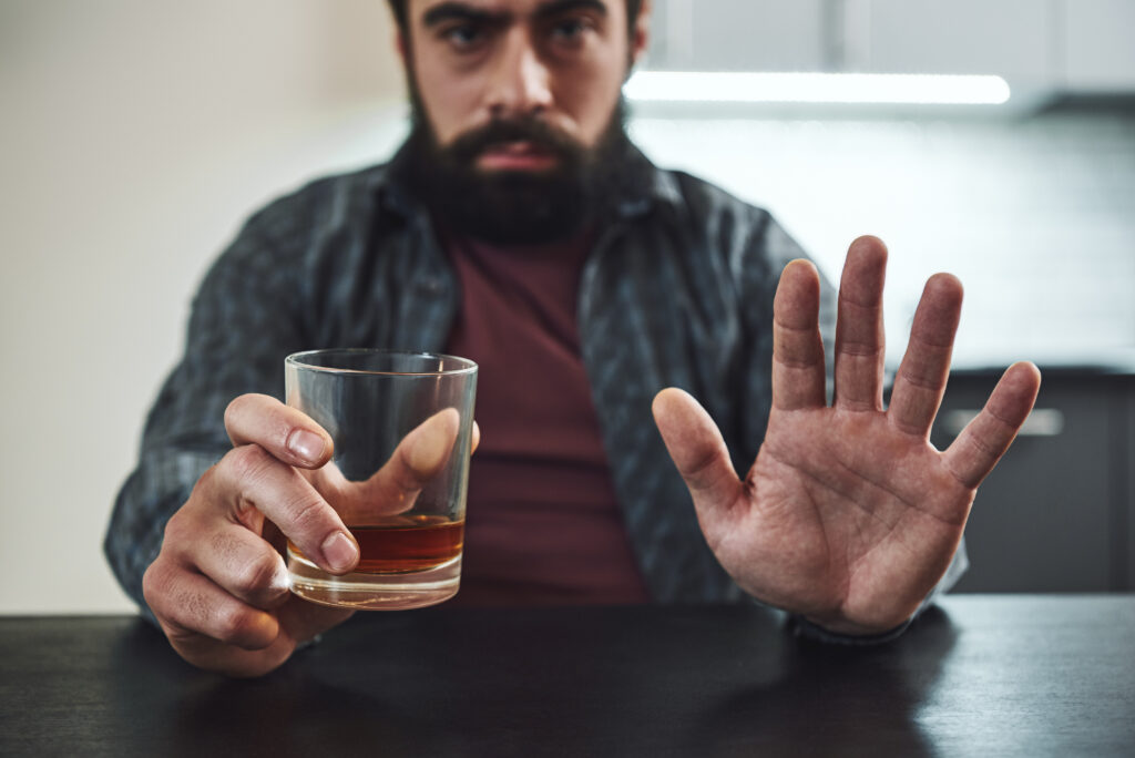 Depressed bearded man sits at the table with a glass of whiskey in his hand. Man refuses to drink alcohol. Male alcoholism concept.