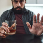 Depressed bearded man sits at the table with a glass of whiskey in his hand. Man refuses to drink alcohol. Male alcoholism concept.