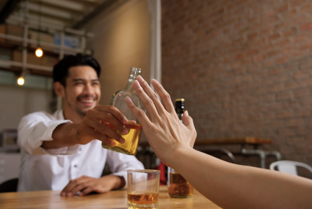 No more alcohol concept. Close up shot of hand refusal alcohol from the man sitting opposite side of table holding bottle of whisky trying to invite him to drink together in night bar restaurant.