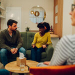 African american woman supporting new attender of group therapy to talk about himself during a meeting at mental health center. Group therapy participants sitting in a circle and talking.