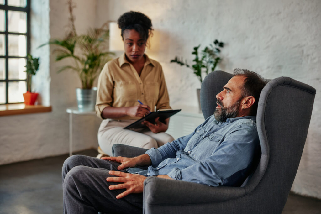 An attentive african female counselor listens and takes notes as a vulnerable middle age male patient discusses an important topic.