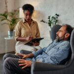 An attentive african female counselor listens and takes notes as a vulnerable middle age male patient discusses an important topic.