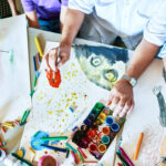 Top view of childrens drawings on table in art class with group of people painting and teachers hands