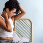 Young multiracial latina woman having neck pain sitting on bed. Female waking up with cervical discomfort. Body concept.