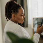 Portrait of good-looking African female student dressed casually holding mobile phone and typing messages and communicating with friends social networks using high-Internet connection.