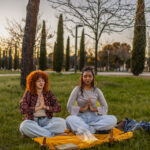 Two multi ethnic female colleagues sitting on a blanket in a park, meditating together after work, embracing the tranquility of the sunset while enjoying nature's beauty