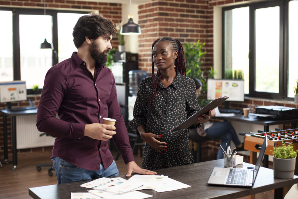 Caucasian businessman holds coffee cup and listens to black woman with pregnancy belly share startup strategy on clipboard. Multiethnic coworkers collaborating on company project in brick wall office.