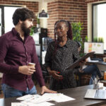 Caucasian businessman holds coffee cup and listens to black woman with pregnancy belly share startup strategy on clipboard. Multiethnic coworkers collaborating on company project in brick wall office.