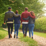 Rear View Of Group Of Young Friends Walking Along Track Through Trees In Countryside Together