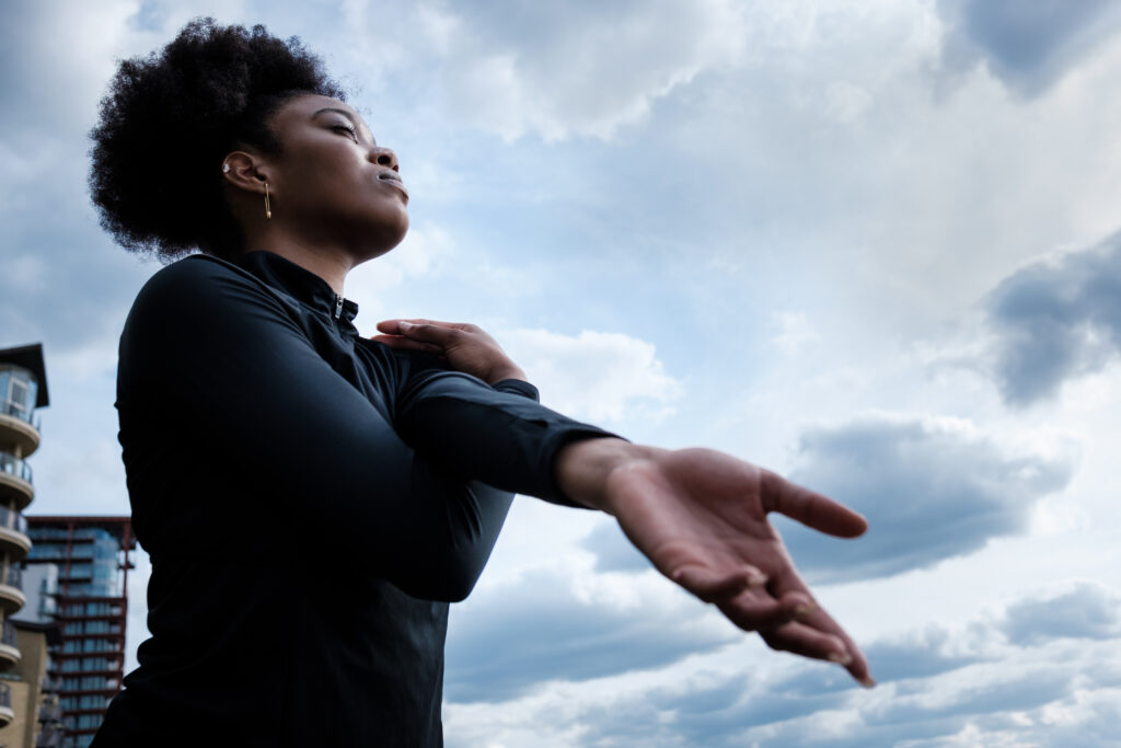Portrait of fit young black woman stretching with a cloudy sky on the background. Curly hair.