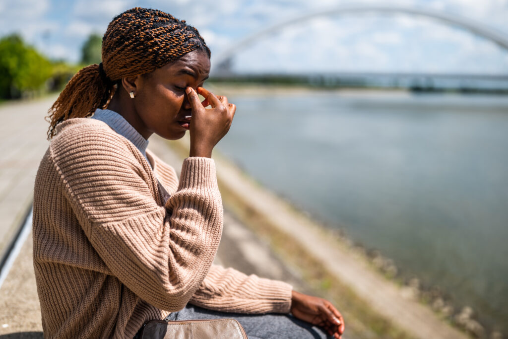 Young beautiful black woman is sitting by the river in the city. She is having headache.