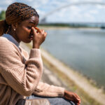 Young beautiful black woman is sitting by the river in the city. She is having headache.