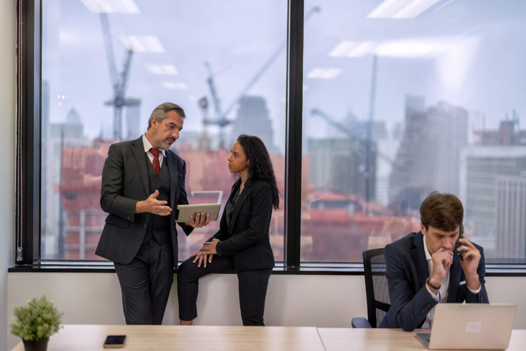 Staff employee sitting use mobile phone call in work place close to window near construction site and overhead crane work in background while colleague talk with manager