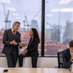 Staff employee sitting use mobile phone call in work place close to window near construction site and overhead crane work in background while colleague talk with manager